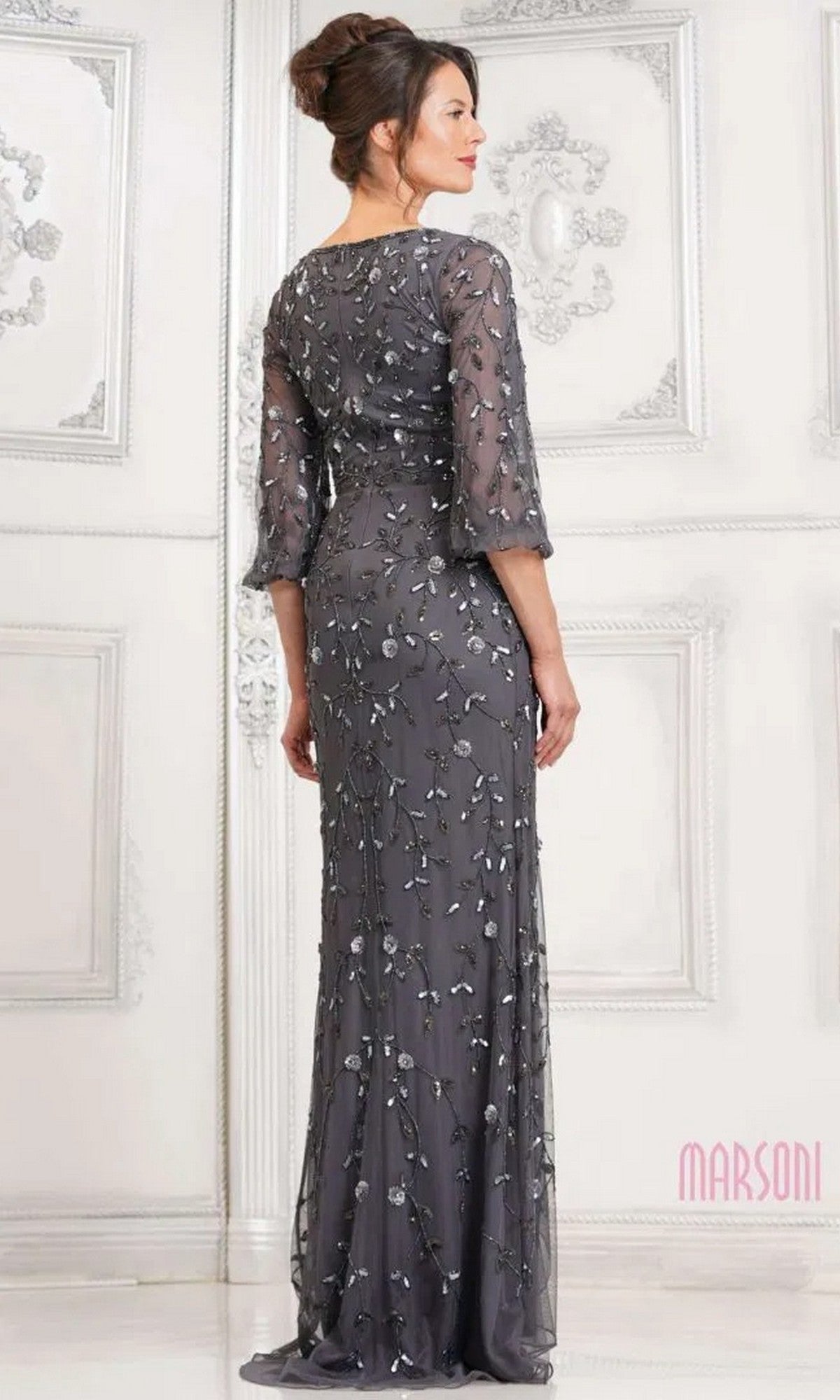 A woman stands facing away in an ornate white room, wearing the Marsoni MV1320 long formal dress—a modest, floor-length dark gray gown with sheer sleeves and silver floral accents.